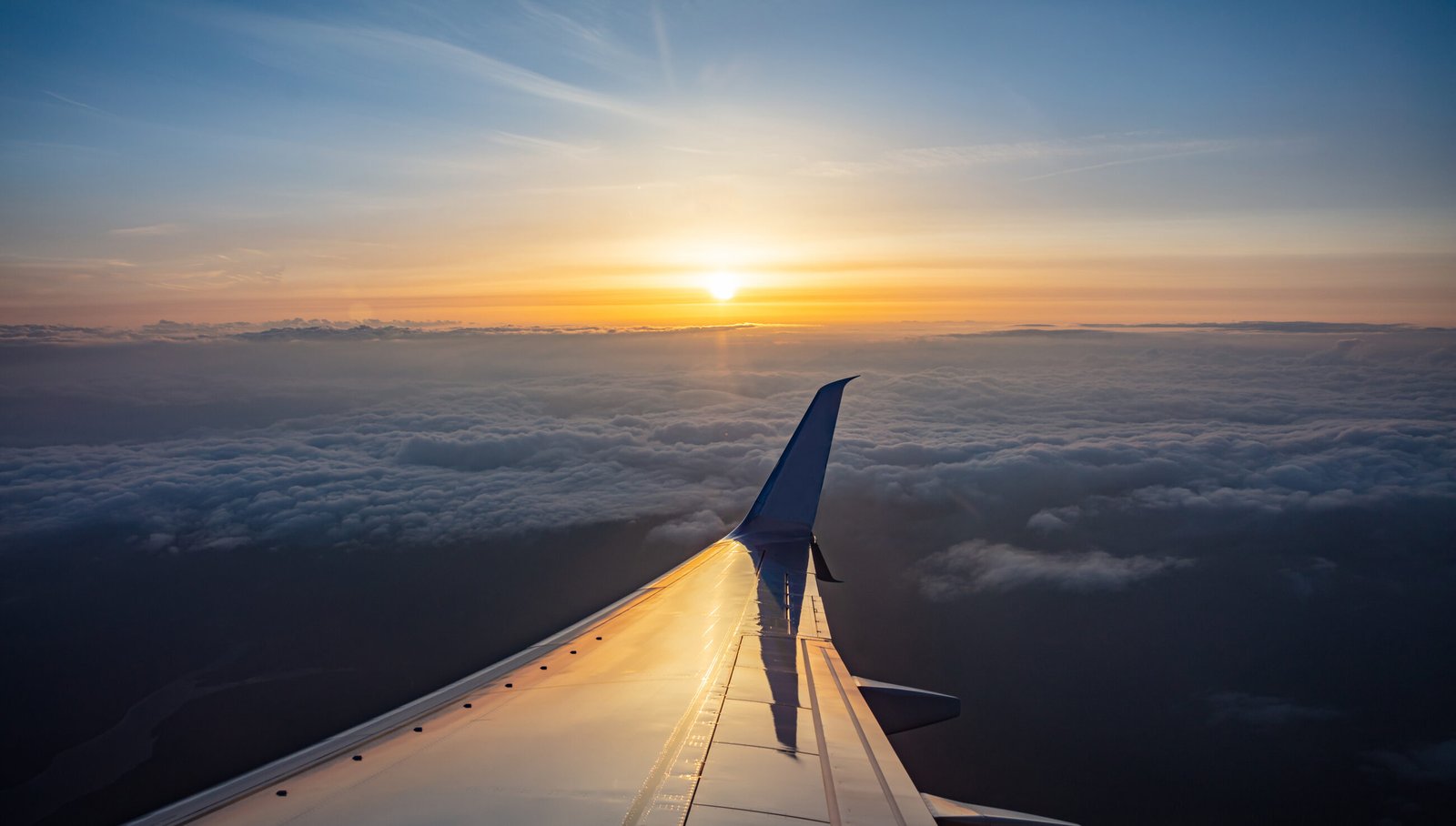 Sunset aerial view out of an airplane window. Sun falling, plane flying over the clouds. Orange color sunbeams and reflections on the plane wing