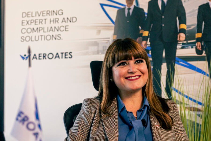 Giovanna Mirabile seated at a desk in an office, smiling at the camera, with an Aeroates aviation display in the background. Workforce Governance in Malta.