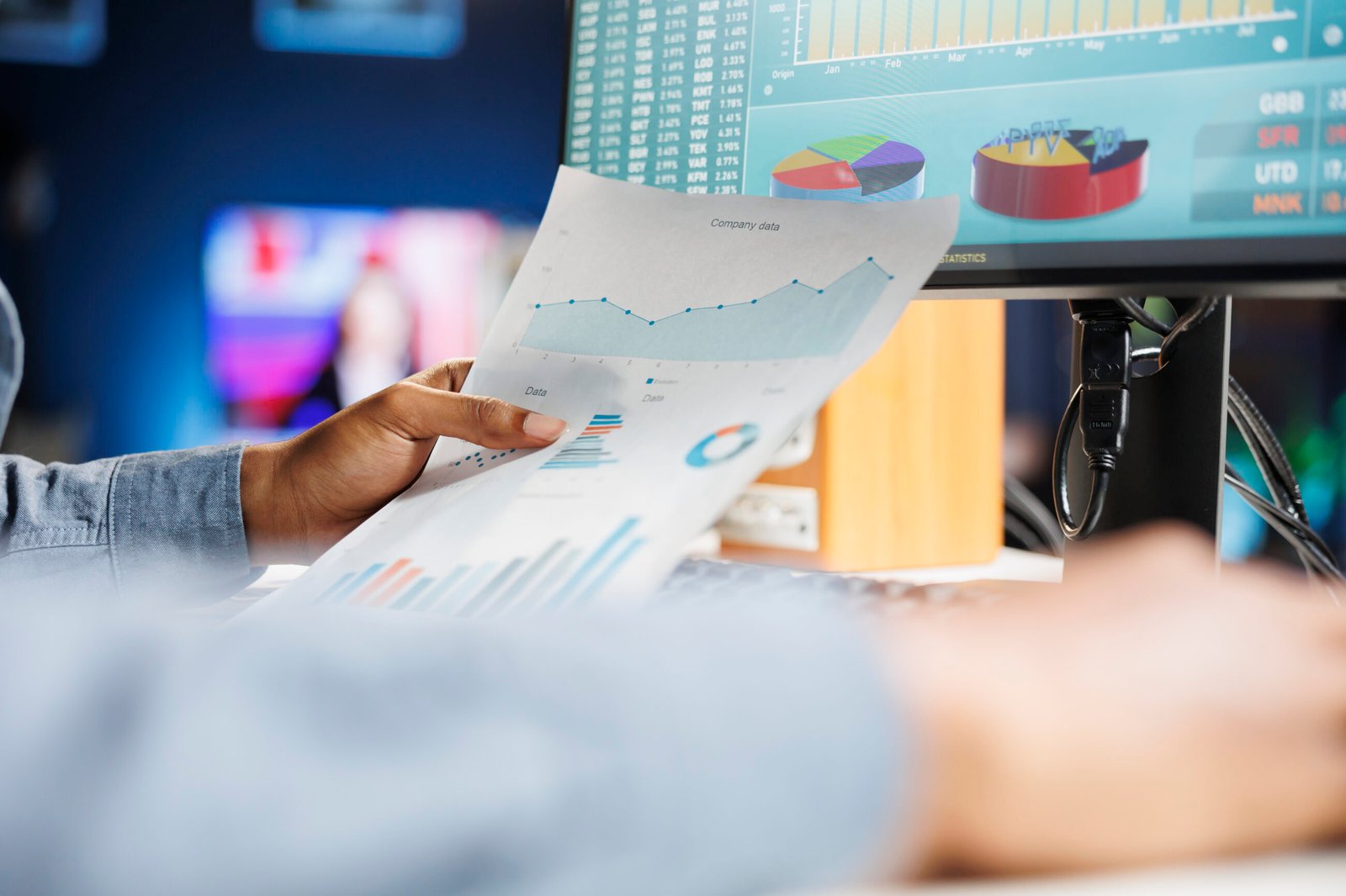 Person reviewing business data charts on paper in front of computer monitors.